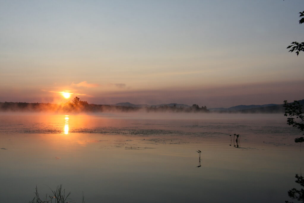 Sunrise over Lake Umbagog with golden light, soft morning fog, calm reflective water, peaceful New England nature photography scene.