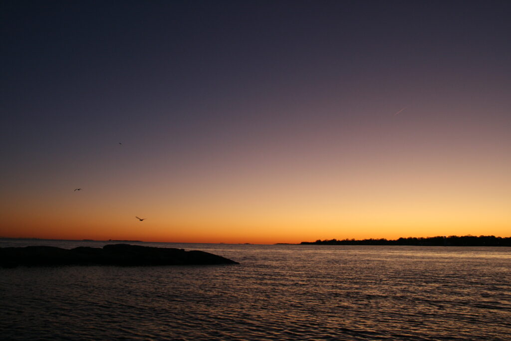 Warm coastal sunset with purple and orange light reflecting on calm water in Noank, Connecticut.