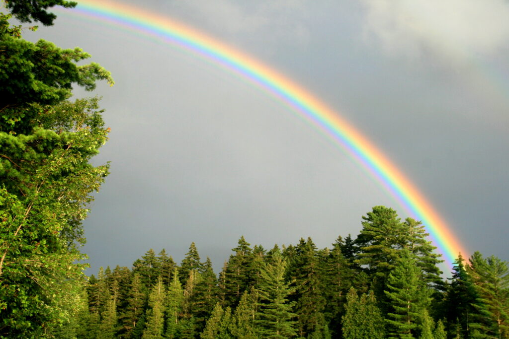 Vibrant rainbow arching over Lake Umbagog with soft light on calm summer water, peaceful New England nature photography.