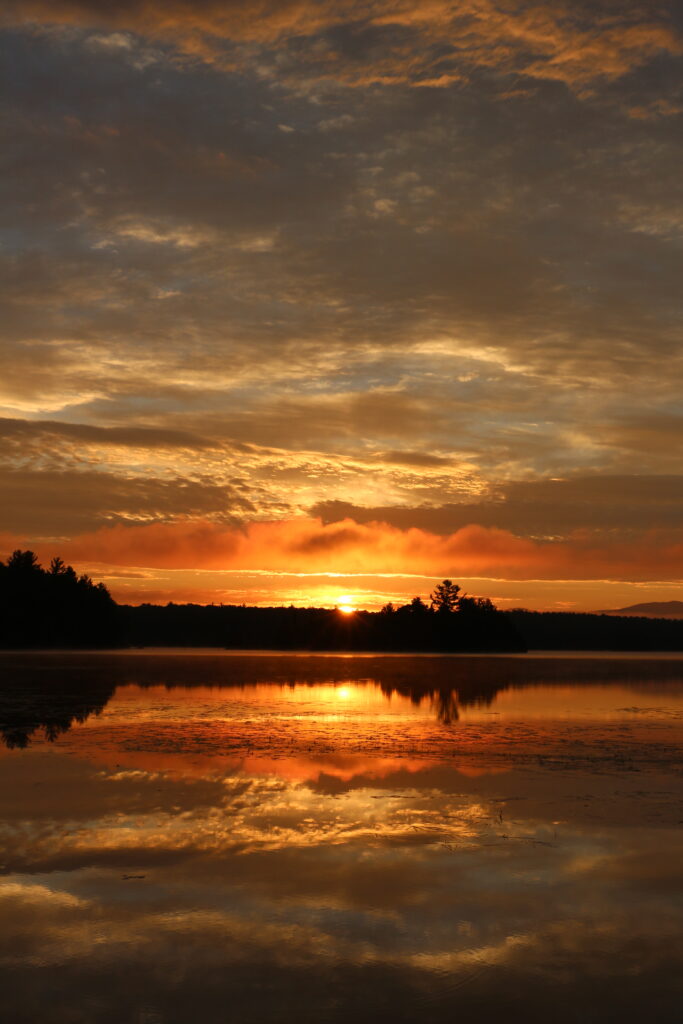 Soft summer sunset with warm orange and gold light reflecting on still water at Lake Umbagog, peaceful New England nature photography.