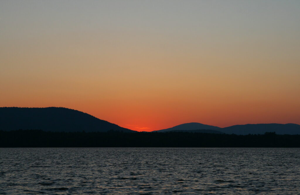 Bands of orange and red sunset light reflecting across calm water at Lake Umbagog, peaceful New England nature photography.