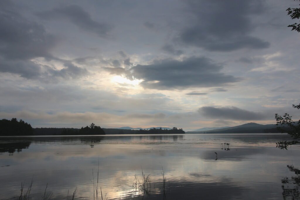 Cloudy sunrise casting soft, muted reflections over the glassy water of Lake Umbagog, with distant mountains and islands along the horizon