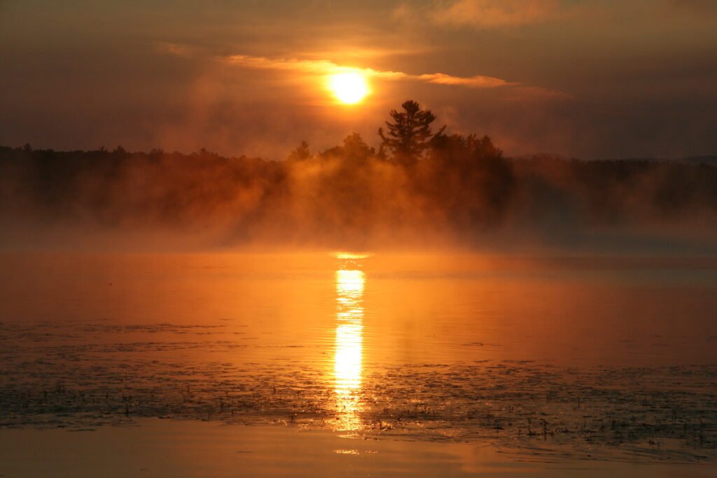 Orange sunrise over Lake Umbagog National Wildlife Refuge with mist, sun rays, and a shadowed island