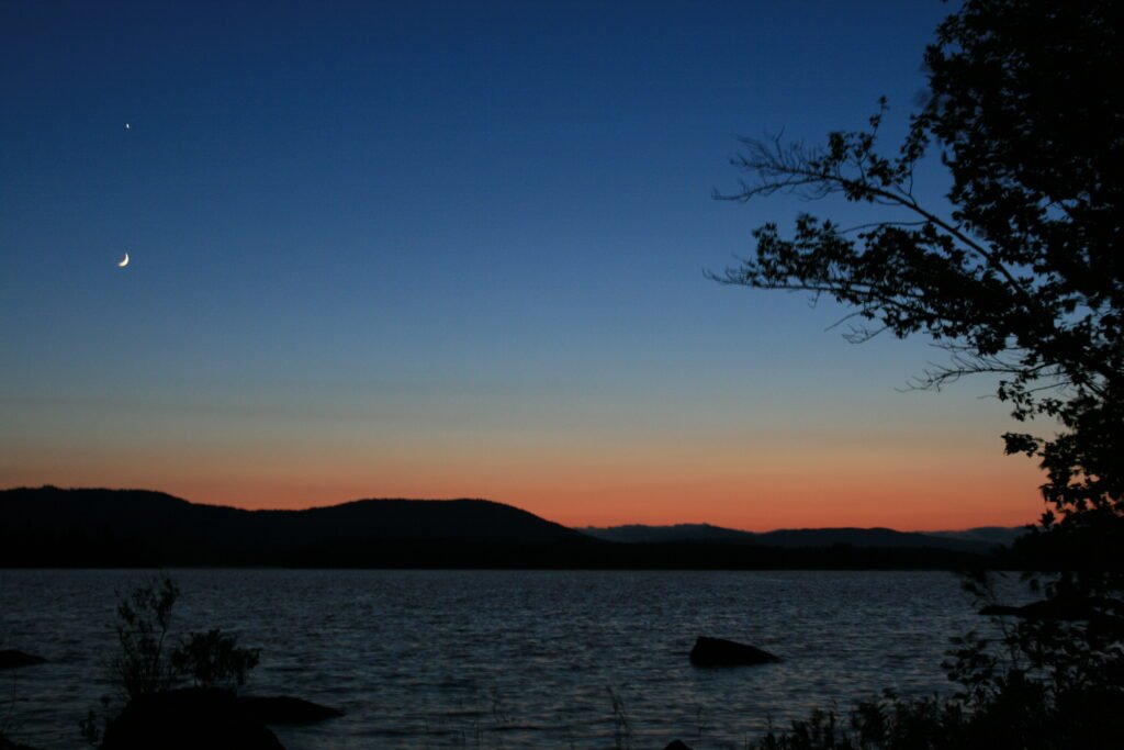 Crescent moon and Venus glowing in the evening twilight above distant mountains and water on Lake Umbagog, New Hampshire